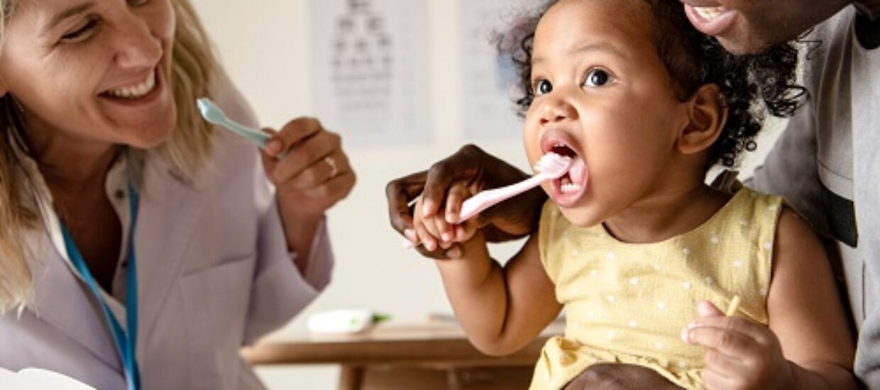 A little girl is in the doctors office with a doctor and caregiver, demonstrating teeth brushing