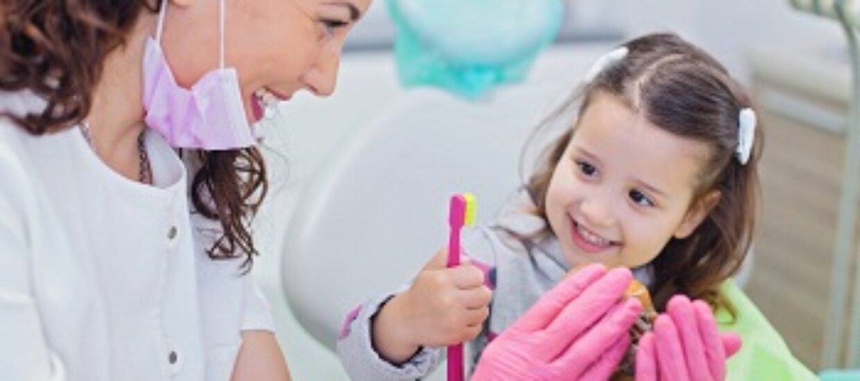 Smiling girl sitting in dentist chair with her pediatric dentist showing her teeth model and teaching her how to brush teeth
