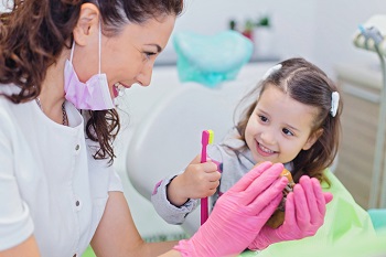 Smiling girl sitting in dentist chair with her pediatric dentist showing her teeth model and teaching her how to brush teeth