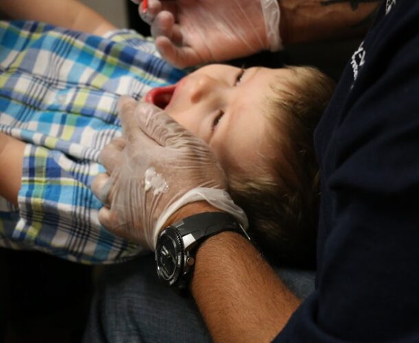 A boy's head is in the lap of a provider who is doing the knee-to-knee exam and painting fluoride varnish on his teeth
