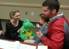 A father with his infant sitting on his lap looking at a woman who is holding a dinosaur puppet.